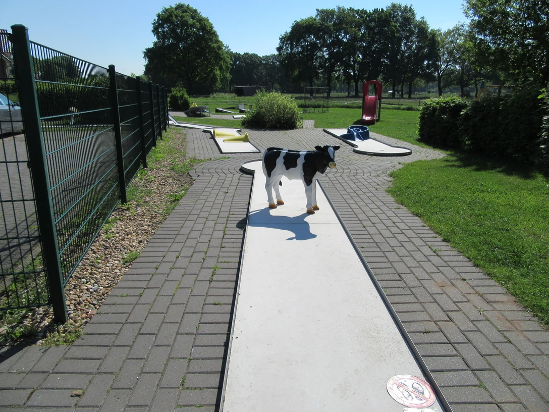 Eine sonnenbeschienene Minigolfbahn mit einem Täuschungsspielzeughund, umgeben von grüner Vegetation.