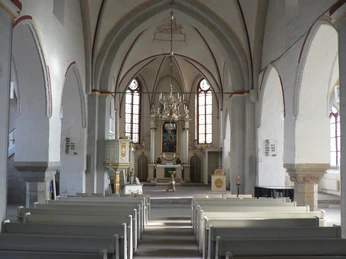 Blick in eine helle Kirche mit weißem Gewölbe, hölzernen Bänken und kunstvollem Altarbild.