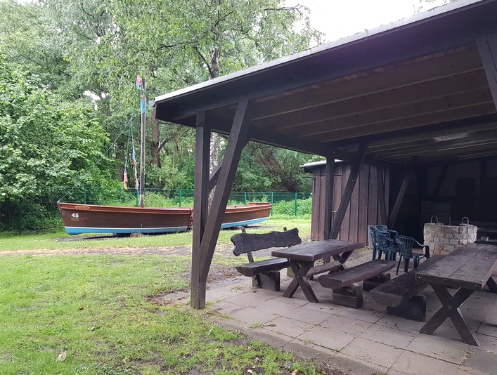 Grillhütte Hagenburg mit Auswanderer Grillhütte aus Holz mit überdachtem Sitzbereich und einem Auswandererschiff im grünen, bewaldeten Hintergrund.