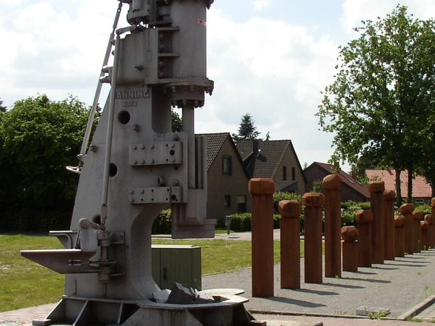 Puddelöfen Eisenhütte Das Bild zeigt eine beeindruckende historische Metallbearbeitungsmaschine, die auf einem gepflasterten Bereich im Freien steht. Im Hintergrund sind Wohnhäuser und Bäume zu sehen, was auf eine ländliche oder kleinstädtische Umgebung hindeutet. Die Maschine, vermutlich ein Puddelofen aus Eisen, besteht aus robustem Metall mit zahlreichen Hebeln und Komponenten, die eine industrielle Funktion andeuten.