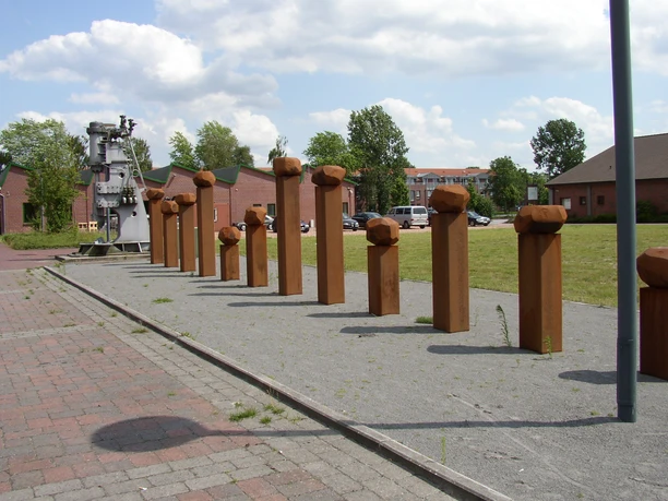 Puddelöfen Eisenhütte Rostrot leuchten hohe, quadratische Säulen mit kugelförmigen Aufsätzen auf einer gepflasterten Fläche. Im Hintergrund sind Ziegelgebäude und Bäume sichtbar, teils verdeckt vom Himmel mit weißen Wolken.