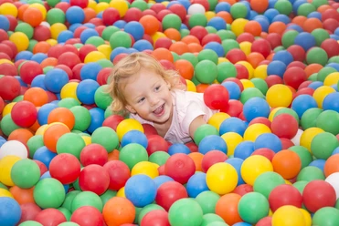Springmaus Kinderspieleland Kind spielt lachend in einem bunten Bällebad mit roten, blauen, gelben, grünen und orangefarbenen Kugeln.