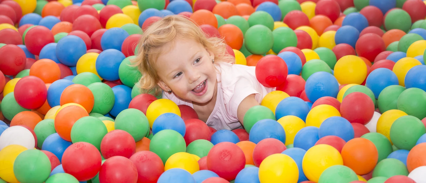 Springmaus Kinderspieleland Kind spielt lachend in einem bunten Bällebad mit roten, blauen, gelben, grünen und orangefarbenen Kugeln.
