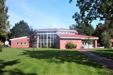 Moderne Freikirche mit rotem Ziegeldach und großem Glasfenster, eingebettet in eine grüne Landschaft.