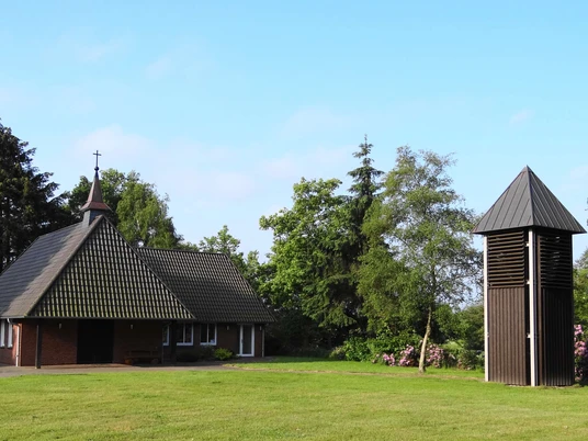 Kleine Kapelle mit dunklem Holzturm, umgeben von grüner Landschaft unter blauem Himmel.