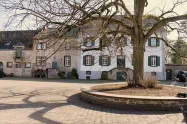 Lindenhof Ein historisches Steingebäude mit grünen Fensterläden und einem weitläufigen, gepflasterten Hof mit einem großen, kahlen Baum im Vordergrund.