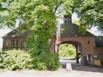 Gut Leidenhaus Historisches Gebäude aus rotem Backstein mit Durchfahrtstor, umgeben von grünen Bäumen.Historic red brick building with drive-through gate, surrounded by green trees.