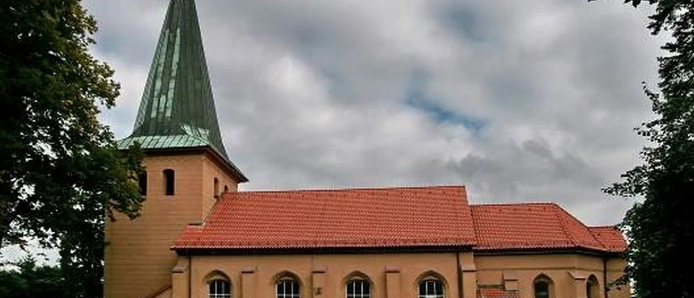Backsteinkirche in Schwarmstedt mit grünem Turm und rotem Dach. Umgeben von Bäumen und Wolken.