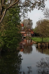 Historische Wassermühle am ruhigen Fluss, umgeben von grünen Bäumen und einem klaren Himmel.