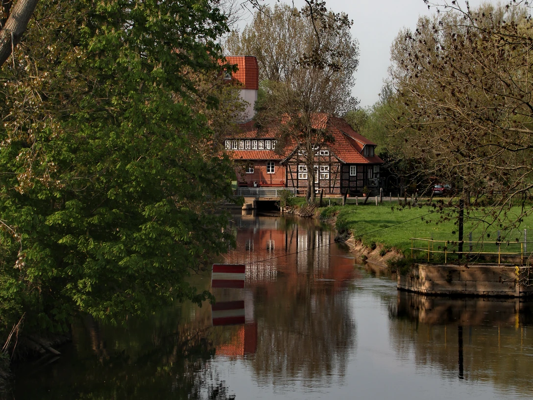 Historische Wassermühle am ruhigen Fluss, umgeben von grünen Bäumen und einem klaren Himmel.