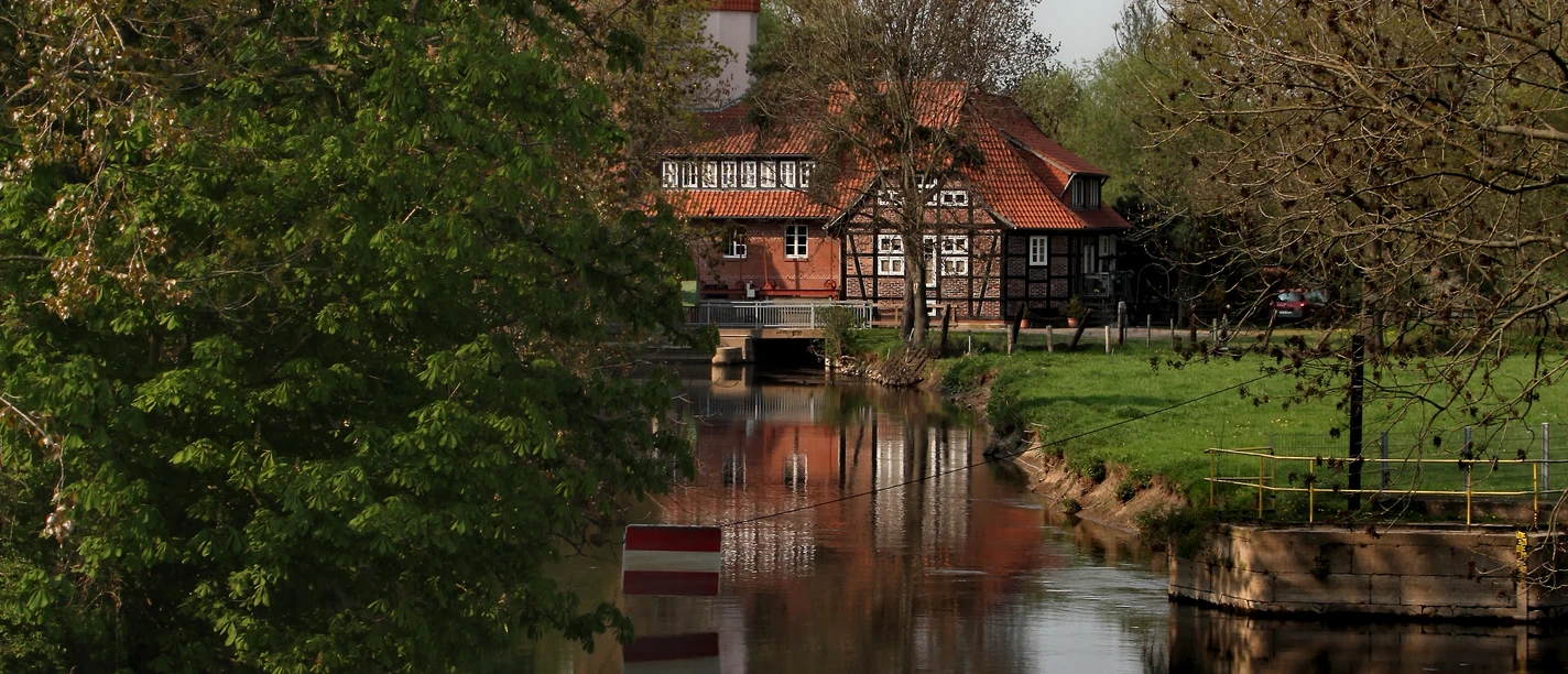 Historische Wassermühle am ruhigen Fluss, umgeben von grünen Bäumen und einem klaren Himmel.