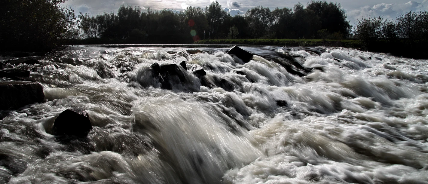 Tosendes Wasser fließt über dunkle Felsen in einer naturnahen Szenerie bei bewölktem Himmel.