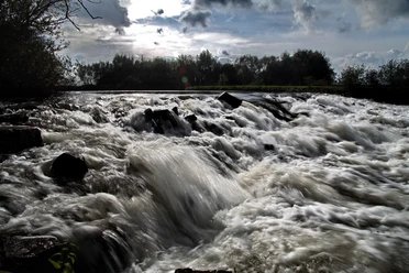 Wasserfall Neustadt a. Rbge. Tosendes Wasser fließt über dunkle Felsen in einer naturnahen Szenerie bei bewölktem Himmel.