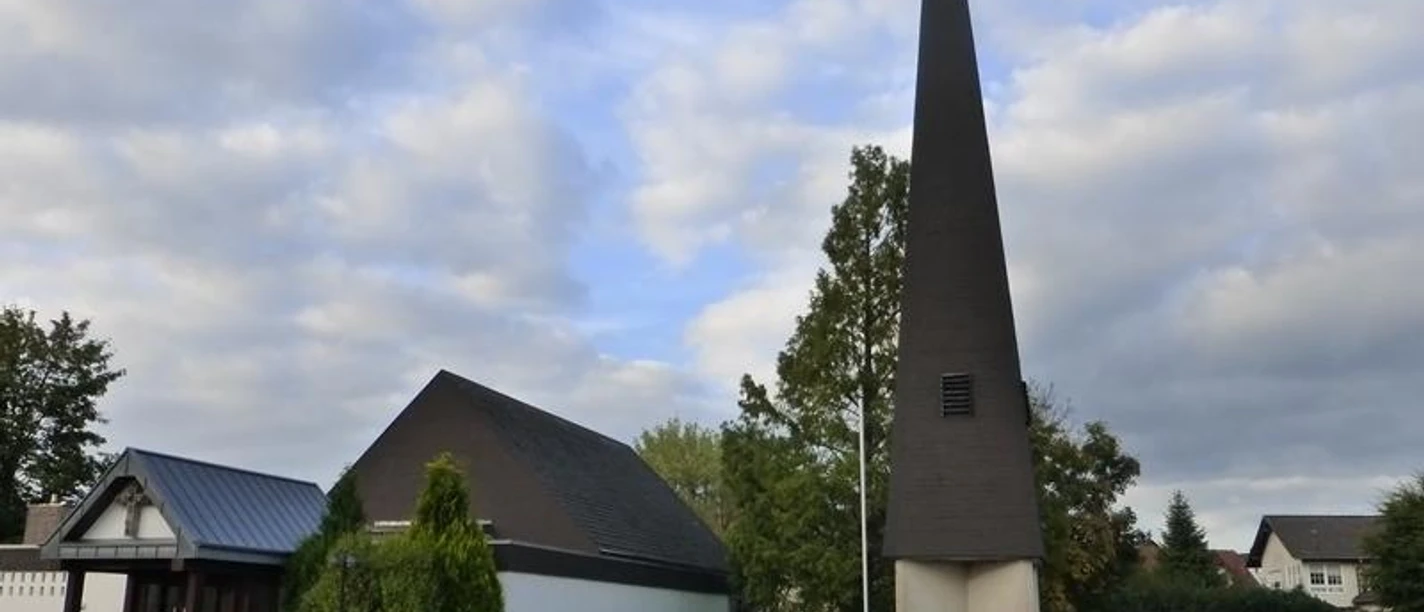 Evangelische Kirche Paderborn-Elsen mit modernem spitzen Turm und umliegenden Bäumen an einem bewölkten Tag.