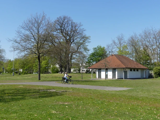Behindertengerechtes WC-Gebäude auf einem offenen Dorfplatz mit Radfahrer und umgebenden Bäumen.