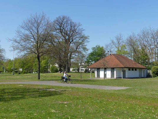 Behindertengerechte WC-Anlage auf dem Dorfplatz Sande Behindertengerechtes WC-Gebäude auf einem offenen Dorfplatz mit Radfahrer und umgebenden Bäumen.