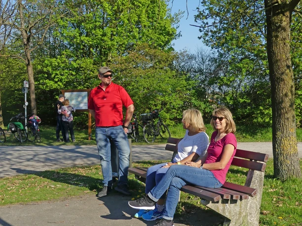 Pause vor der Tour. Im Hintergrund die Infotafel der WasserRoute. Drei Personen entspannen auf einer Parkbank nahe einer Infotafel zur WasserRoute, Fahrräder im Hintergrund.