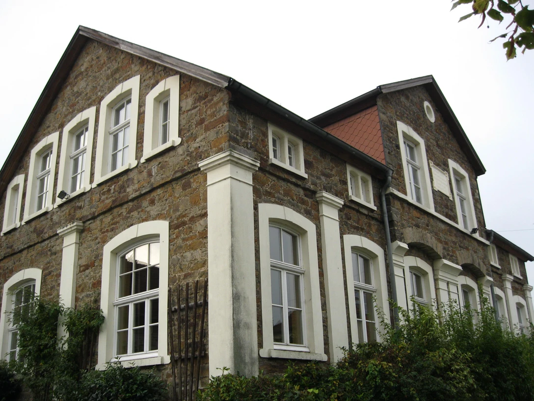 Alte Schule Almena Historisches Steingebäude mit weißen Fensterrahmen und begrüntem Vorgarten unter bewölktem Himmel.