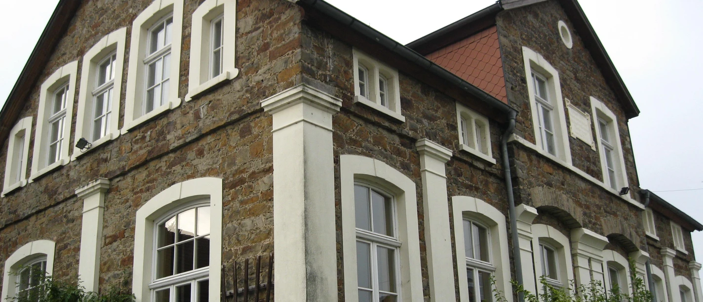 Alte Schule Almena Historisches Steingebäude mit weißen Fensterrahmen und begrüntem Vorgarten unter bewölktem Himmel.