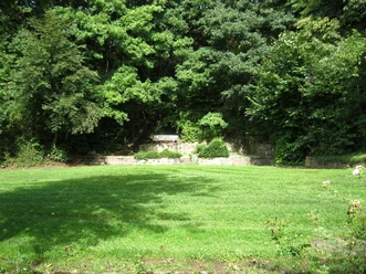 Der Rosenbergpark in Extertal-Almena Grüner Rasen umgeben von Bäumen, eine Parkbank steht vor einer steinernen Wand im Rosenbergpark.