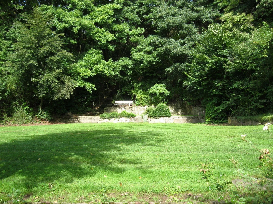 Der Rosenbergpark in Extertal-Almena Grüner Rasen umgeben von Bäumen, eine Parkbank steht vor einer steinernen Wand im Rosenbergpark.