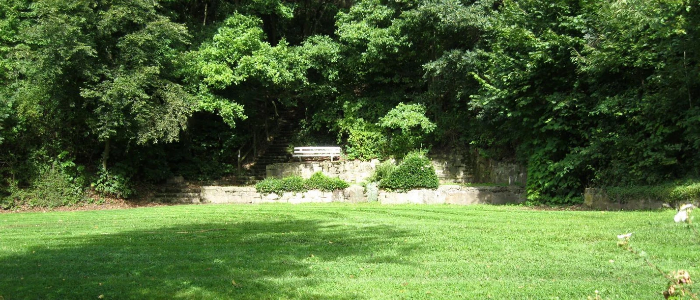 Der Rosenbergpark in Extertal-Almena Grüner Rasen umgeben von Bäumen, eine Parkbank steht vor einer steinernen Wand im Rosenbergpark.