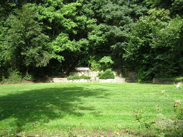 Der Rosenbergpark in Extertal-Almena Grüner Rasen umgeben von Bäumen, eine Parkbank steht vor einer steinernen Wand im Rosenbergpark.