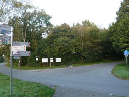 Parkplatz am Boker Kanal Schloß Neuhaus: asphaltierte Fläche mit Wegweisern und bewaldeter Kulisse.