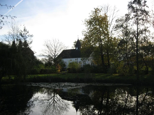 Alte Schule mit Dorfteich und Parkanlage Eine historische Schule spiegelt sich malerisch in einem ruhigen Teich, umgeben von Bäumen.