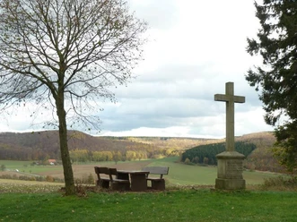 Gedenkkreuz und Picknickbank neben einem kahlen Baum, mit Blick auf weite, hügelige Landschaft.