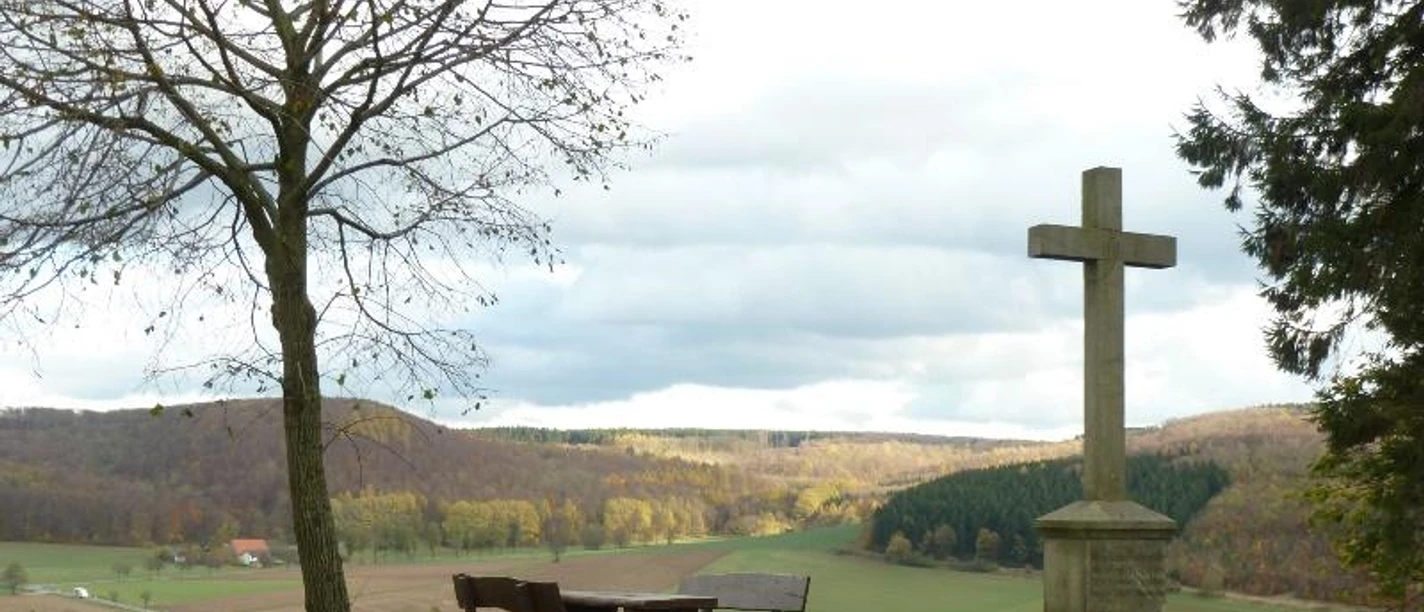 Gedenkkreuz und Picknickbank neben einem kahlen Baum, mit Blick auf weite, hügelige Landschaft.