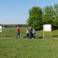 Menschen spazieren auf einem Lehrpfad entlang Infotafeln, umgeben von einer weitläufigen, grünen Landschaft.