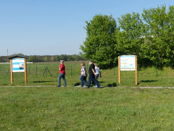 Menschen spazieren auf einem Lehrpfad entlang Infotafeln, umgeben von einer weitläufigen, grünen Landschaft.