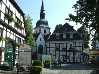 Fachwerkkirche mit Turm, umgeben von historischen Fachwerkhäusern und einem offenen Platz bei Sonnenschein.