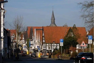 Fachwerkhäuser mit roten Dächern, ein Kirchturm, geparkte Autos und Straßenlaternen.