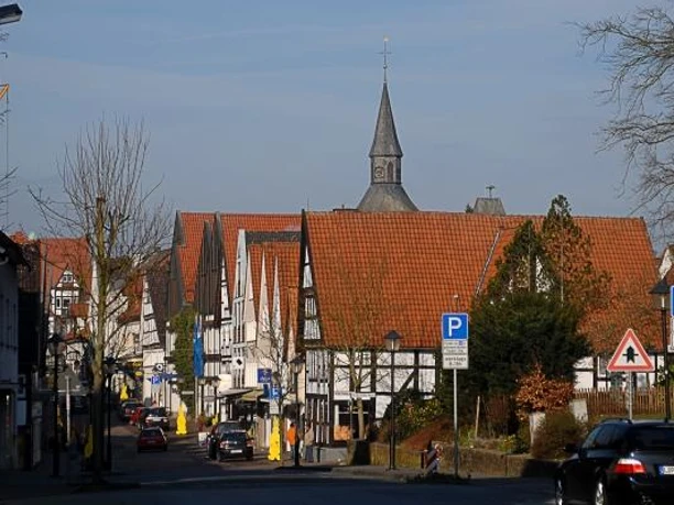 Fachwerkhäuser mit roten Dächern, ein Kirchturm, geparkte Autos und Straßenlaternen.
