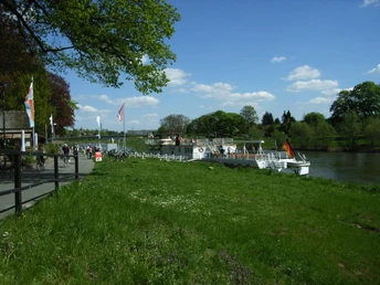 Grünes Ufer am Schiffsanleger Höxter mit Ausflugsboot, Spazierweg und blühenden Bäumen im Hintergrund.