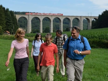 Fünf Wanderer vor einem imposanten Viadukt, über das ein roter Zug fährt, umgeben von Natur.