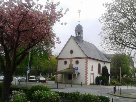 Eine kleine Kapelle mit Schindeldach und Turm, umgeben von blühenden Bäumen und Straßenpflaster.