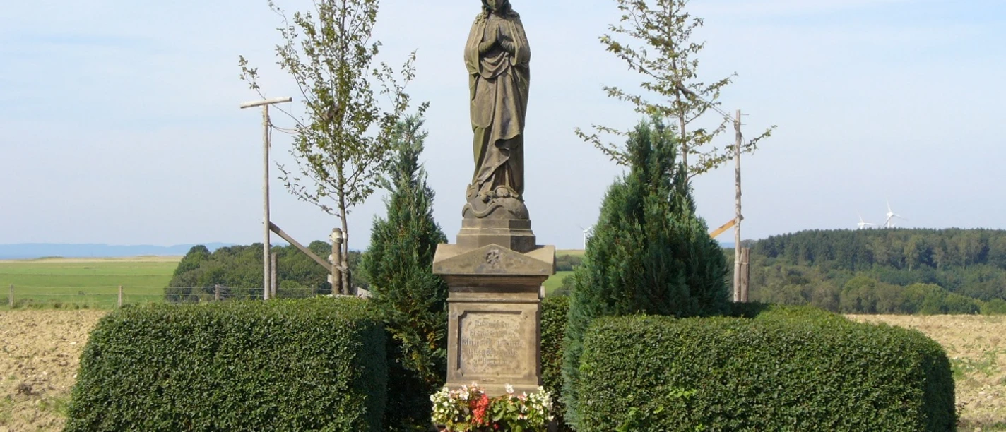 Madonnenstandbild auf einem Feld bei Paderborn-Dahl, umgeben von Bäumen und gepflegten Büschen.