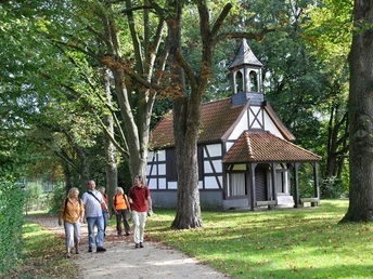 Fachwerk-Kapelle mit Glockenturm, umgeben von Bäumen; vier Menschen spazieren auf einem Weg davor.