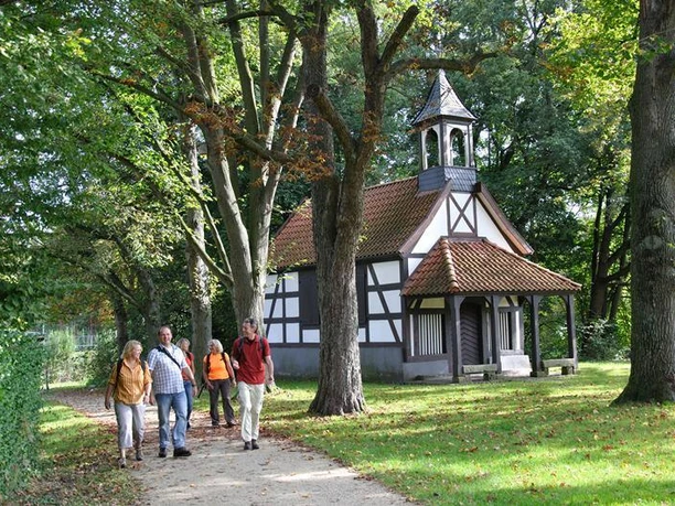 Rochuskapelle Fachwerk-Kapelle mit Glockenturm, umgeben von Bäumen; vier Menschen spazieren auf einem Weg davor.