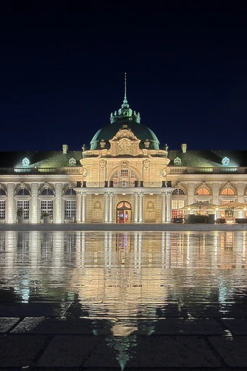 GOP Kaiserpalais Bad Oeynhausen Aussenansicht Das GOP Kaiserpalais Bad Oeynhausen, majestätisch erleuchtet bei Nacht, spiegelt sich auf einem nassen Platz.