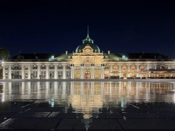 GOP Kaiserpalais Bad Oeynhausen Aussenansicht Das GOP Kaiserpalais Bad Oeynhausen, majestätisch erleuchtet bei Nacht, spiegelt sich auf einem nassen Platz.