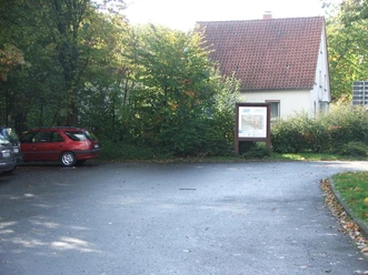 Parkplatz im Ortskern mit asphaltierten Stellflächen, angrenzend an ein Gebäude und umgeben von Bäumen.