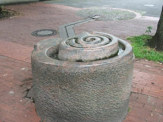 Brunnenanlage "Der Weg" Rund geformter Brunnen aus Stein mit spiralförmigem Design, auf einem gepflasterten Platz gelegen.