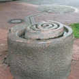 Brunnenanlage "Der Weg" Rund geformter Brunnen aus Stein mit spiralförmigem Design, auf einem gepflasterten Platz gelegen.