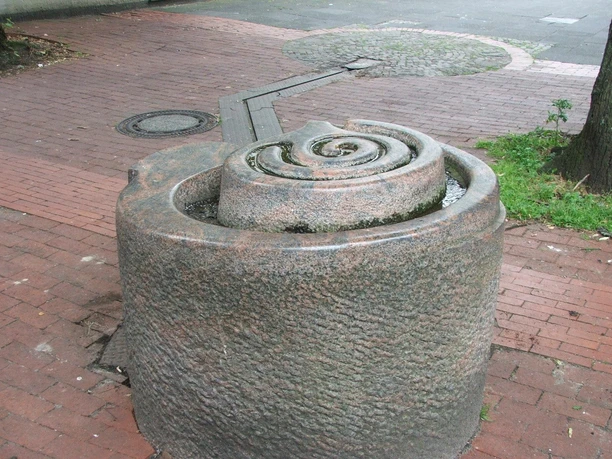 Brunnenanlage "Der Weg" Rund geformter Brunnen aus Stein mit spiralförmigem Design, auf einem gepflasterten Platz gelegen.