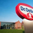 Dr. Oetker-Firmenschild vor einem modernen Gebäude mit historischen Backstein-Elementen und blauen Himmel.