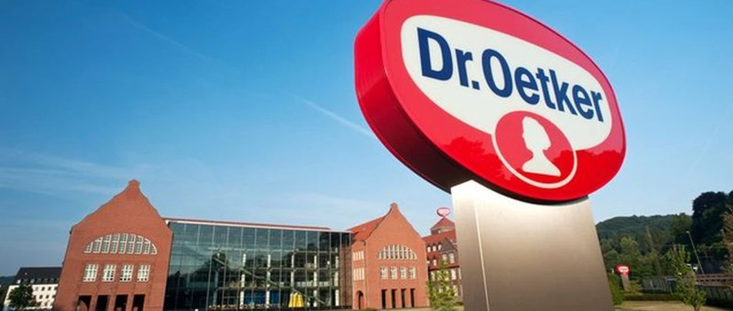 Dr. Oetker Dr. Oetker-Firmenschild vor einem modernen Gebäude mit historischen Backstein-Elementen und blauen Himmel.
